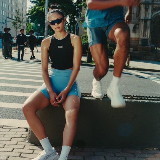 Stamina Luna Race Crop in black, modeled on a woman sitting with a man running nearby, urban setting.