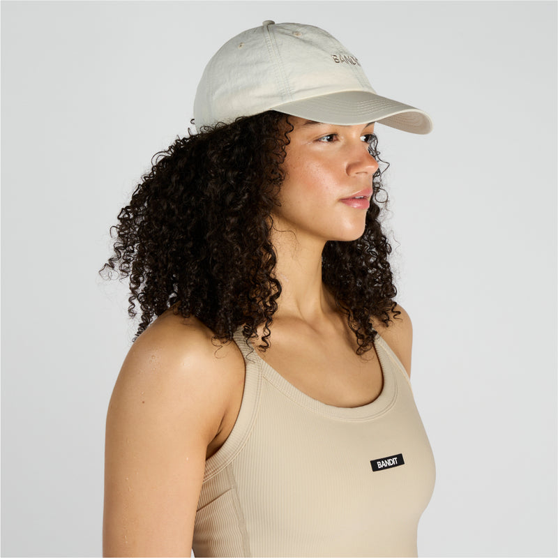 Cream nylon dad hat with embroidered wordmark, featuring adjustable webbing and clip detail, worn by a model.