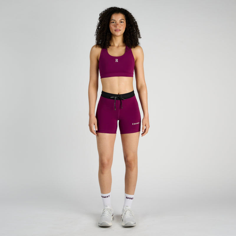 Stamina™ 5" women's compression shorts in plum, worn by model, featuring dual side pockets, zippered back pocket, and custom waistband.